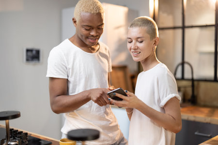 Multiracial couple using mobile phone together at home kitchen. Concept of relationship. Idea of modern domestic lifestyle. Black man and european girl spending time together. Interior of modern flatの写真素材
