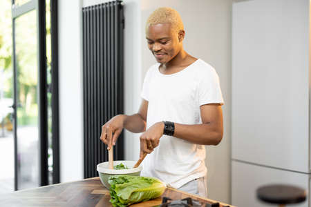 Black blonde man cooking organic salad at home kitchen. Idea of healthy eating. Concept of modern domestic lifestyle. Smiling handsome guy standing at table. Interior of modern apartmentの写真素材