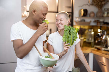 Multiracial couple eating organic salad at home kitchen. Idea of healthy eating. Concept of relationship. Modern domestic lifestyle. Black man and european girl spending time togetherの写真素材
