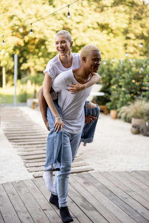 Black boyfriend giving piggyback ride for his caucasian girlfriend on home terrace. Concept of relationship and enjoying time together. Modern domestic lifestyle. Warm sunny daytime.の写真素材