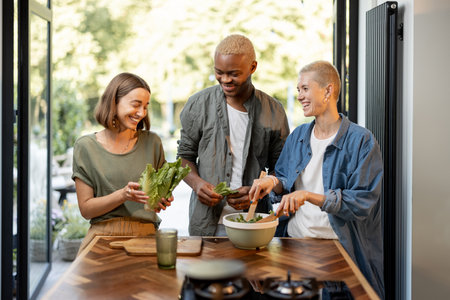 Friends cooking salad at home kitchen. Black man and european girls enjoying time together. Concept of healthy eating. Modern lifestyle friendship. Interior of apartment. Sunny daytimeの写真素材