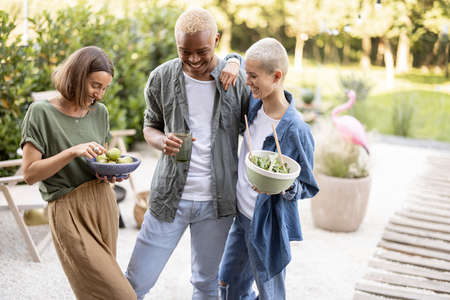 Friends enjoying time together at home backyard. Black man and european girls with organic food. Concept of healthy eating. Idea of friendship. Modern lifestyle. Summer sunny daytimeの写真素材