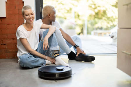 Multiracial couple sitting on floor at home with robot vacuum cleaner. European girl and black man spending time together. Concept of modern domestic lifestyle. Idea of house cleaningの写真素材