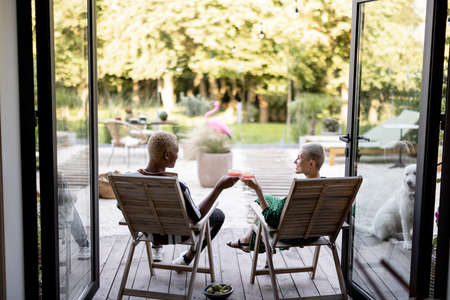 Multiracial couple drinking cocktails while sitting on wooden chairs at home terrace. European girl and black man spending time together. Concept of leisure. Modern domestic lifestyleの写真素材