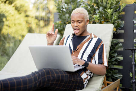 Black man using laptop computer while laying on lounger at house garden. Concept of leisure. Idea of modern domestic lifestyle. Young excited guy having new idea. Summer sunny daytimeの写真素材