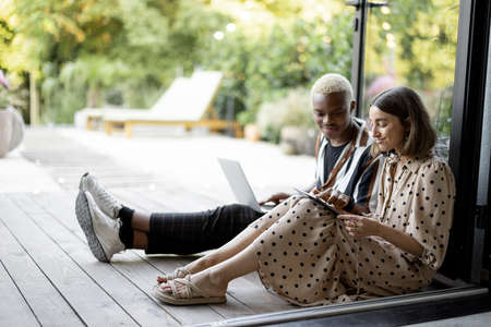 Black man with laptop watching something on digital tablet of his european girlfriend. Couple on wooden terrace at home. Concept of leisure and spending time together. Modern domestic lifestyleの写真素材