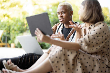 European girl showing something on digital tablet to her focused black boyfriend with laptop. Couple on wooden terrace at home. Concept of leisure and spending time together. Modern domestic lifestyleの写真素材