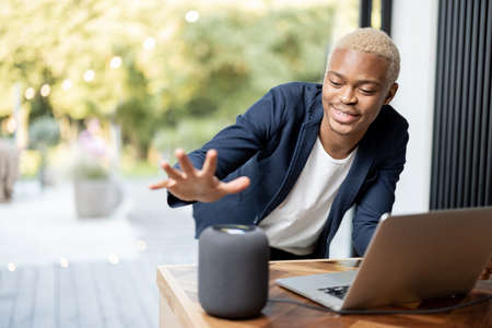 Black man taking portable speaker from wooden table with laptop computer. Concept of modern lifestyle. Idea of audio technology. Young smiling handsome guy. Interior of sunny day in modern apartment.の写真素材