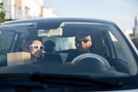 Multiracial couple riding together in car. Young european woman holding groceries after shopping. Concept of relationship. Black man and girl in glasses. Modern lifestyle. Sunny daytimeの写真素材