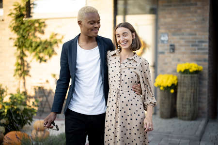 Multiracial couple going together on background of their house. Concept of relationship and enjoying time together. Young black man embracing his caucasian girlfriend. Modern lifestyle. Sunny daytimeの写真素材