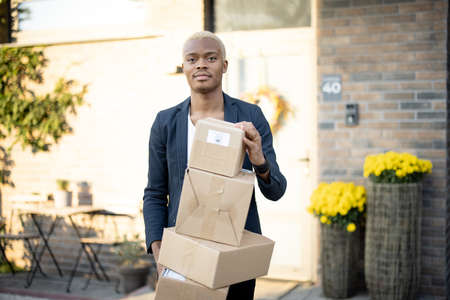 Blonde black man standing with parcels with background of his house. Young serious guy holding cardboard boxes and looking at camera. Modern lifestyle. Sunny daytime. Concept of housewarming.の写真素材