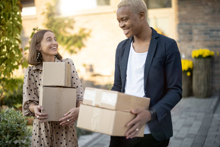 Multiracial couple walking with parcels with background of their house. Young smiling caucasian woman and black man holding cardboard boxes outdoors. Modern lifestyle. Autumn sunny daytime.の写真素材