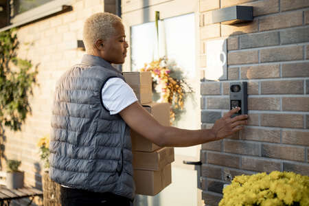 Courier with parcels ringing in doorbell of client`s house. Concept of shipping and logistics. Young blonde black guy in gray waistcoat is a worker of delivery service. Autumn sunny daytime.の写真素材