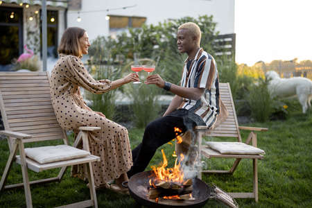 Multiracial couple drinking and toasting cocktails during home party in garden. Burning fire for bbq. Concept of relationship. Domestic lifestyle. Black man and european girl enjoying time togetherの写真素材