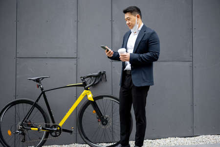 Asian businessman using mobile app on smartphone for bicycle rent on city street. Concept of modern transportation. Idea of online application. Man with coffee cup wearing suitの写真素材