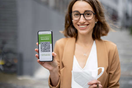 Caucasian businesswoman holding medical mask and showing smartphone with app of Coronavirus Vaccination on city street. Concept of health protection during pandemic. Joyful young woman in glassesの写真素材