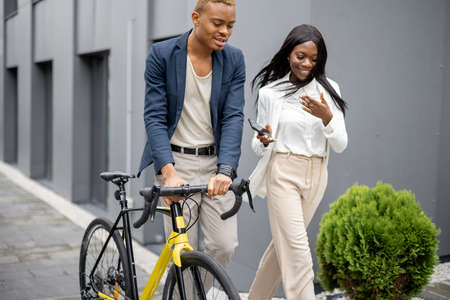 Businessman and businesswoman walking on city street and talking. Concept of remote and freelance work. Idea of business cooperation. Young man with bicycle. Beautiful woman with smartphoneの写真素材