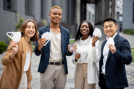 Multiracial business people holding medical masks and showing smartphone with app of Coronavirus Vaccination on city street. Concept of health protection during pandemic. Joyful young businessteamの写真素材
