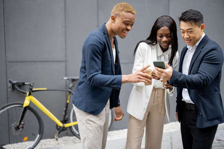 Multiracial business team watching something on smartphone on city street. Concept of break on job. Idea of team building. Smiling businesswoman and businessmen wearing formal wearの写真素材