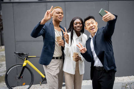 Multiracial business team showing victory gesture while taking selfie on smartphone. Concept of break on job. Idea of team building. Smiling businesswoman and businessmen on city streetの写真素材