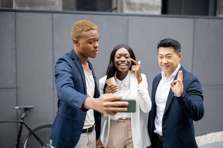 Multiracial business team taking selfie on smartphone on city street. Concept of break on job. Idea of team building. Joyful businesswoman and businessmen wearing formal wearの写真素材