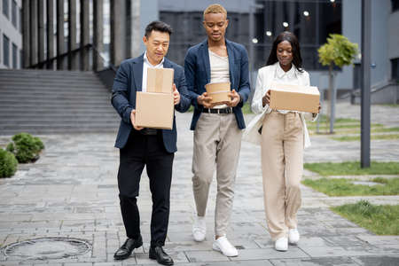 Multiracial business team with parcels and food walking on city street. Concept of remote and freelance work. Idea of shipping and logistics. Focused businesswoman and businessmen wearing formal wearの写真素材