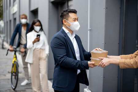 Businessman taking food order from unidentified woman who working in closed cafe. Concept of business during Coronavirus pandemic. Multiracial businesspeople in medical masks waiting in queueの写真素材