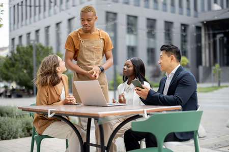Waiter taking order from clients at outdoor cafe. Multiracial business team having lunch during break on work. Concept of teambuilding and corporate event. Businesspeople sitting at table with gadgetsの写真素材