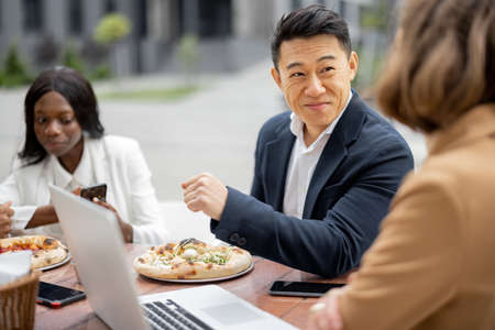 Multiracial businessteam having lunch with pizza and talking at outdoor cafe. Concept of teambuilding and corporate event. Idea of rest and leisure on job. People at table with digital devicesの写真素材
