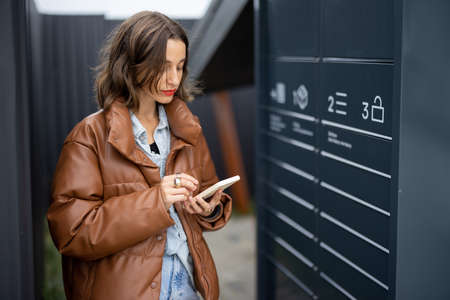 Young caucasian woman using smartphone near automatic post terminal. Focused girl on city street. Concept of smart delivery. Idea of modern shipping and logisticsの写真素材