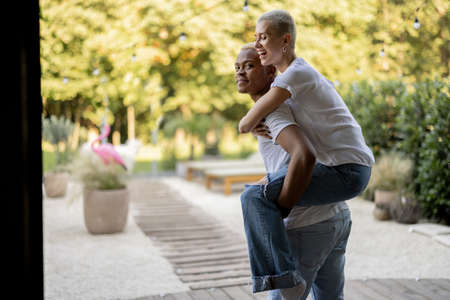 Latin boyfriend giving piggyback ride for his caucasian girlfriend on home terrace. Concept of relationship and enjoying time together. Modern domestic lifestyle. Warm sunny daytime.の写真素材