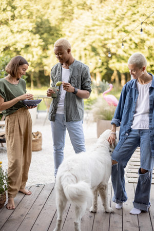 Friends enjoying time with a dog together at home backyard. Black man and european girls with organic food. Concept of healthy eating. Idea of friendship. Modern lifestyle. Summer sunny daytimeの写真素材
