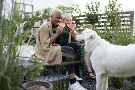 Multiracial couple eating watermelon in their garden. Concept of relationship and enjoying time together with a dog. Black man hugging his european girlfriend. People covered in plaidの写真素材