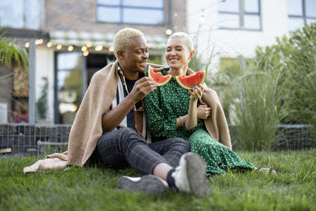 Multiracial couple eating watermelon in their garden. Concept of relationship and enjoying time together. Modern lifestyle. Black man hugging his european girlfriend. People covered in plaidの写真素材
