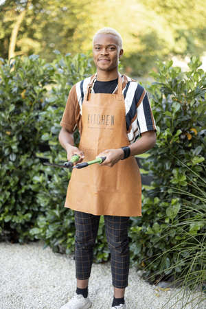Portrait of a Latin man cutting green plant with gardening scissors in his garden. Concept of summer leisure. Idea of modern domestic lifestyle. Young focused handsome guy wearing apronの写真素材