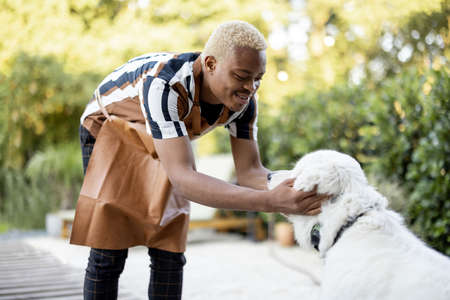 Black man playing with a dog. Concept of gardening and leisure. Young smiling handsome guy wearing apron. Idea of modern domestic lifestyle on summer sunny daytime.の写真素材