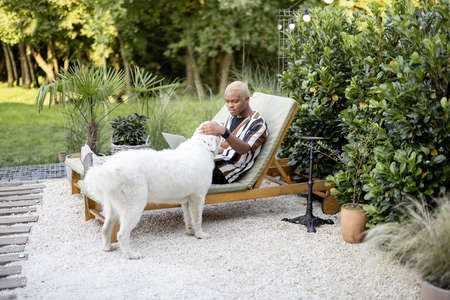 Latin man playing with a dog while laying and resting on lounger at house garden. Concept of leisure. Idea of modern domestic lifestyle. Young focused guy. Summer sunny daytimeの写真素材
