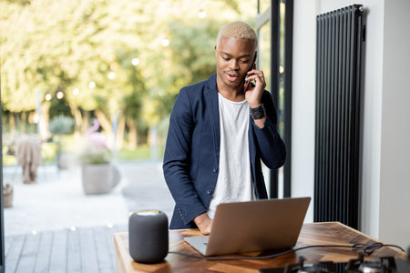 Latin man talking on phone while working on laptop. Controlling smart home devices remotely with a voice command. Concept of smart home. Idea of audio technologyの写真素材