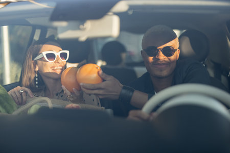 Multiracial couple riding together in car. Young european woman holding organic groceries after shopping. Concept of relationship. Black man and girl in glasses enjoy time together. Modern lifestyleの写真素材