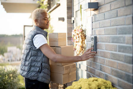 Courier with parcels ringing in doorbell of client s house. Concept of shipping and logistics. Young blonde black guy in gray waistcoat is a worker of delivery service. Autumn sunny daytimeの写真素材