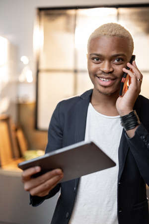 Latin man using digital tablet while talk on smartphone. Concept of modern domestic lifestyle. Young focused guy work remotely from home. Interior of kitchen in modern apartmentの写真素材