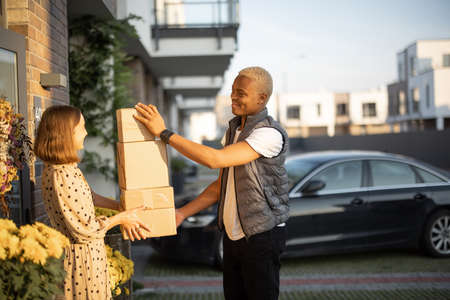 Delivery man giving parcels to caucasian woman on entrance of her house. Young smiling girl taking cardboard boxes. Concept of shipping and logistics. Idea of delivery serviceの写真素材