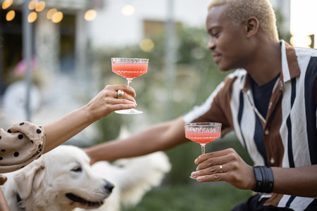 Multiracial couple drinking cocktails and caressing dog during home party in their garden. Burning fire for bbq. Concept of relationship rest. Black guy and european girl enjoying time together.の写真素材