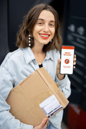 Young caucasian woman holding parcel and showing smartphone with smart delivery app. Smiling girl near automatic post terminal in city. Concept of modern shipping and logisticsの写真素材