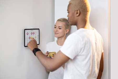 Latin man controlling smart home devices on a digital tablet, while his european girlfriend looking at him. Concept of modern domestic technologies. Couple looking at each other in kitchen.の写真素材