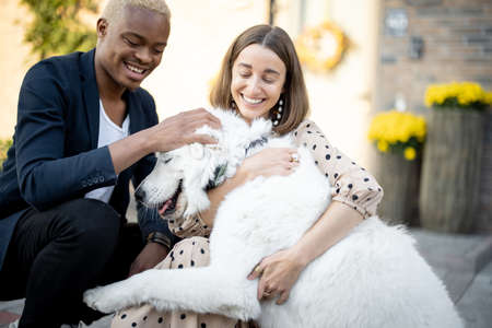 Multiracial couple caress dog on background of thier house. Concept of relationship and enjoying time together. Young smiling caucasian girl and black man. Modern lifestyle. Autumn sunny daytimeの写真素材