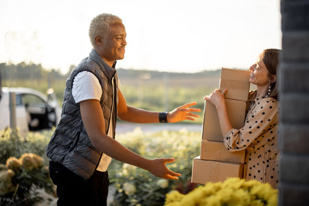 Delivery man giving parcels to caucasian woman on entrance of her house. Young smiling girl taking cardboard boxes. Concept of shipping and logistics. Idea of delivery serviceの写真素材