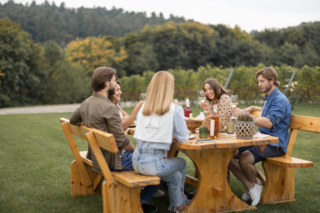 Joyful european friends drinking and toasting with local wine during picnic near vineyards in countryside. Young men and women spend time together. Concept of winemaking. Friendship and leisure.の写真素材
