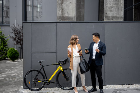 Asian businessman and caucasian businesswoman drinking coffee and using smartphones on city street...