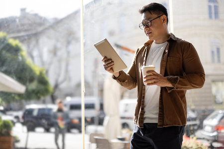 Asian businessman drinking coffee and watching something on digital tablet inside unknown building....